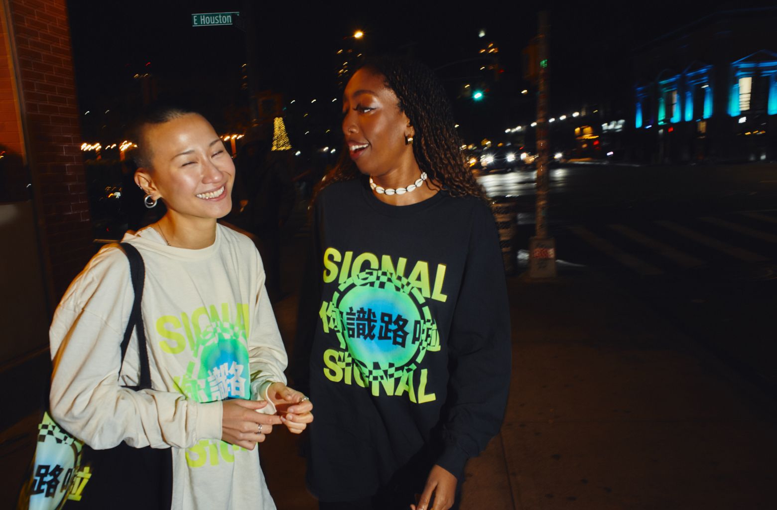 Two friends walking down the street at night wearing Signal longsleeve shirts and holding a Signal tote bag.