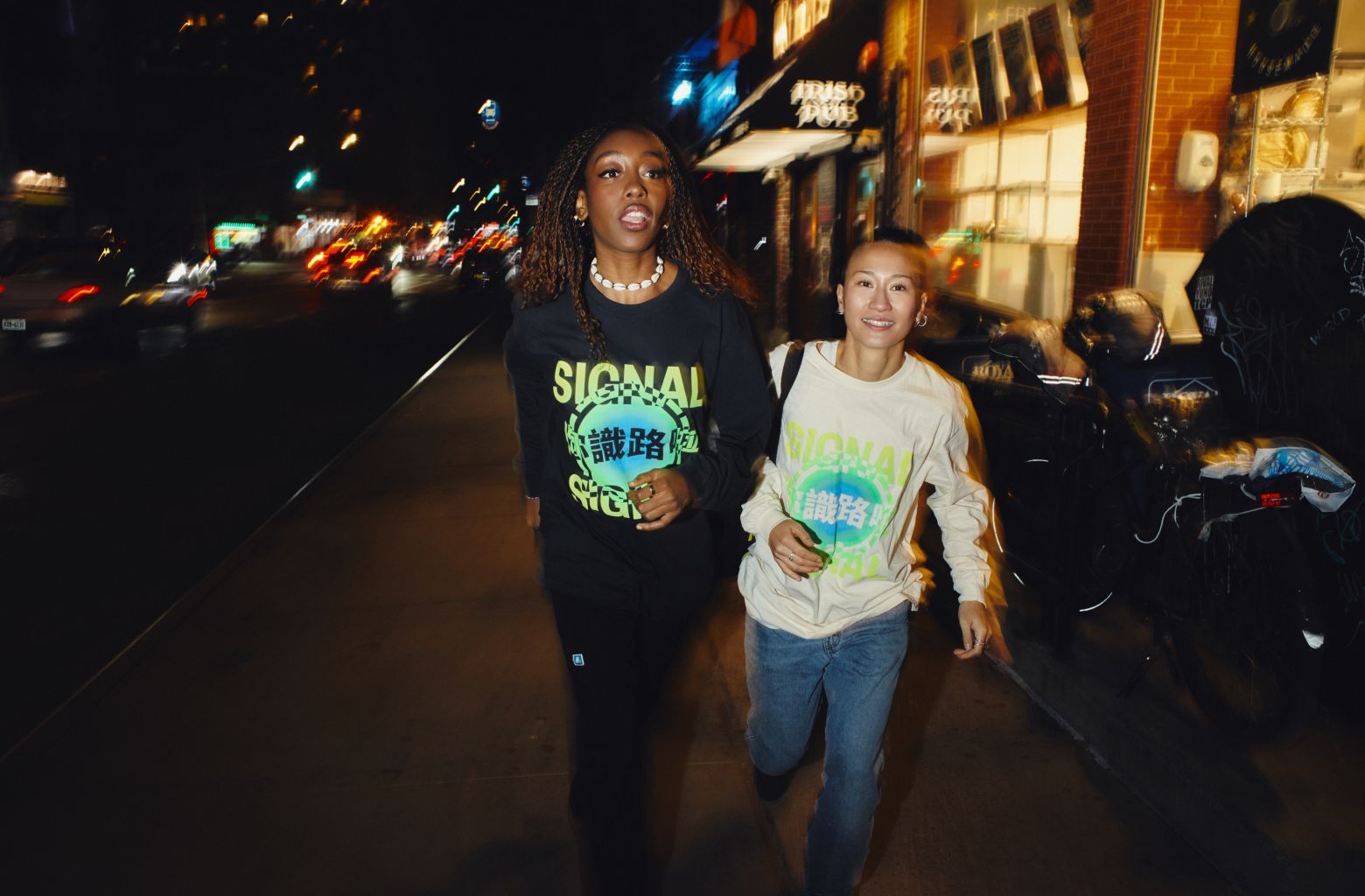 Two friends laughing on the street at night wearing Signal longsleeve shirts and holding a Signal tote bag.